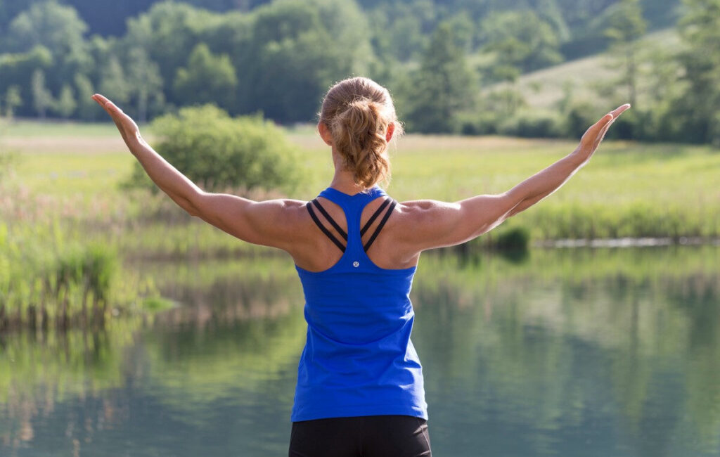 yoga-am-see-valerie-froehlich Blonde Frau mit Pferdeschwanz von hinten vor einem See mit grüner Umgebung im Sommer. Sie hebt die durchtrainierten Arme. Die Sonne scheint.