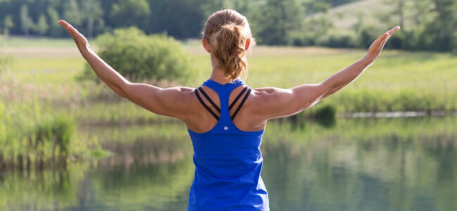 yoga-am-see-valerie-froehlich Blonde Frau mit Pferdeschwanz von hinten vor einem See mit grüner Umgebung im Sommer. Sie hebt die durchtrainierten Arme. Die Sonne scheint.